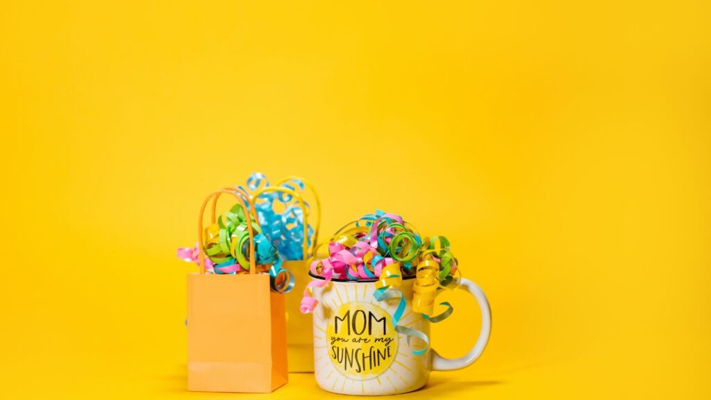 Mother's Day gifts - a small orange paper gift bag and a coffee mug that reads "Mom, you are my sunshine" - against a bright yellow background