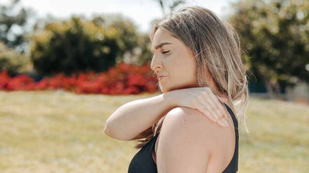 A young woman in athletic wear walking outdoors rubs her shoulder as if in pain